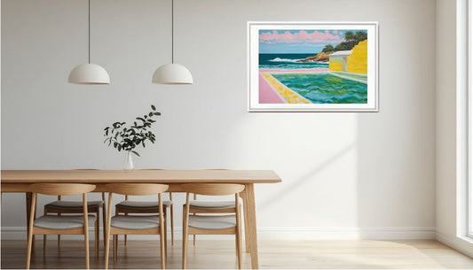 Framed colourful artwork of a seascape with a yellow house and pool hung above a dining table in a room with white walls and pendant lights.