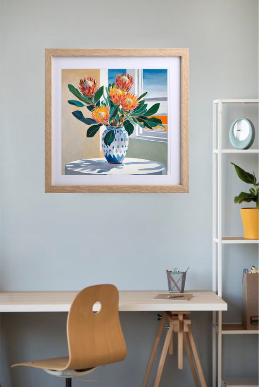 orange and pink proteas in blue and white vase sitting on white table near window art print framed in wood frame hanging on wall