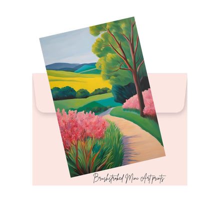 Landscape mini print with pink flowers and green trees  hills and a path on top of a pink envelope, featuring 'Brushstroked mini Art Prints' branding.