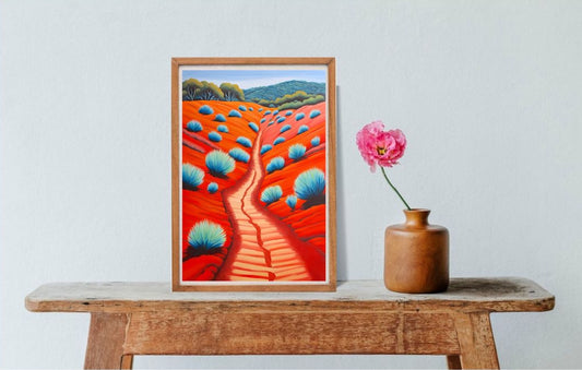 Framed artwork of an Australian landscape on a wooden table with a vase and pink flower against a white wall.